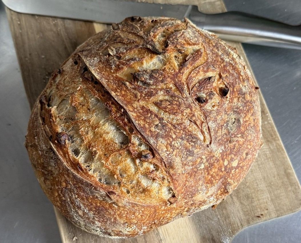 Walnut and pecan sourdough boule with a deeply browned crust, dusted with flour and scored with a leaf pattern, resting on a wooden peel with a bread knife behind it