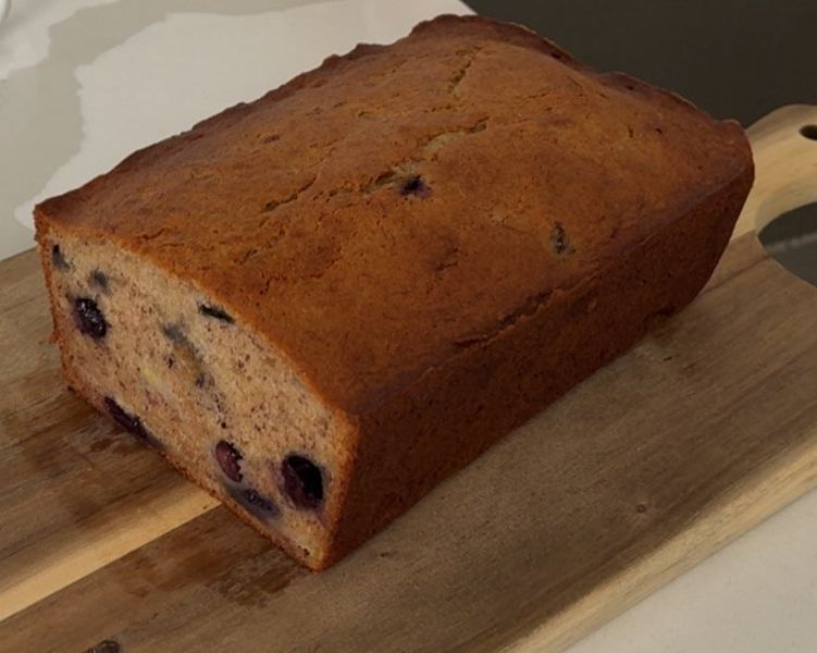 Blueberry banana bread loaf on a wooden cutting board with a thick slice cut from one end, whole blueberries speckled through the golden crumb