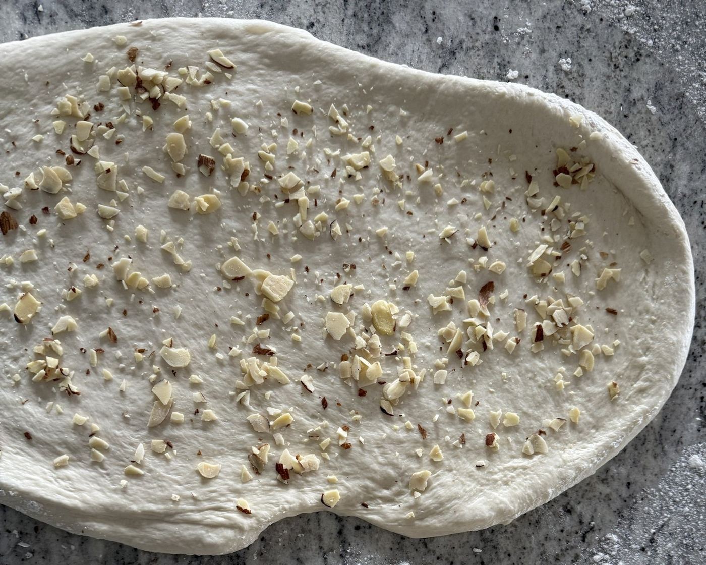 Stretched dough sprinkled with chopped almonds on a granite counter beside a rolling pin