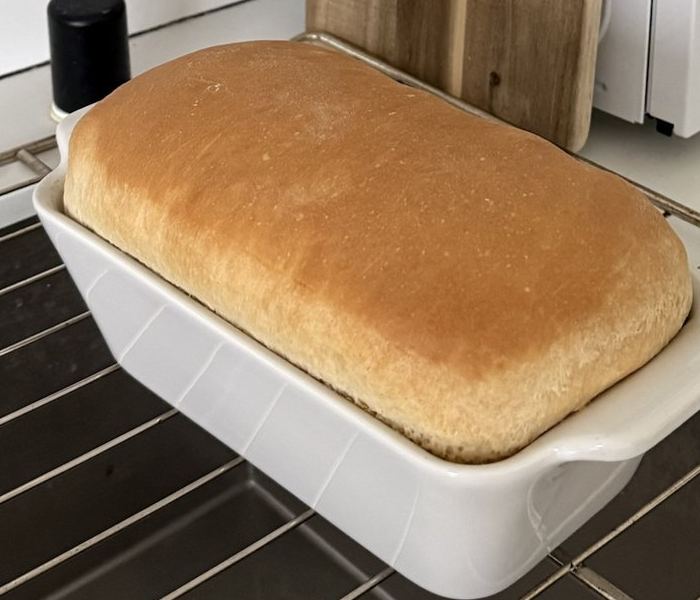 Freshly baked white bread still in its white ceramic loaf pan on a cooling rack