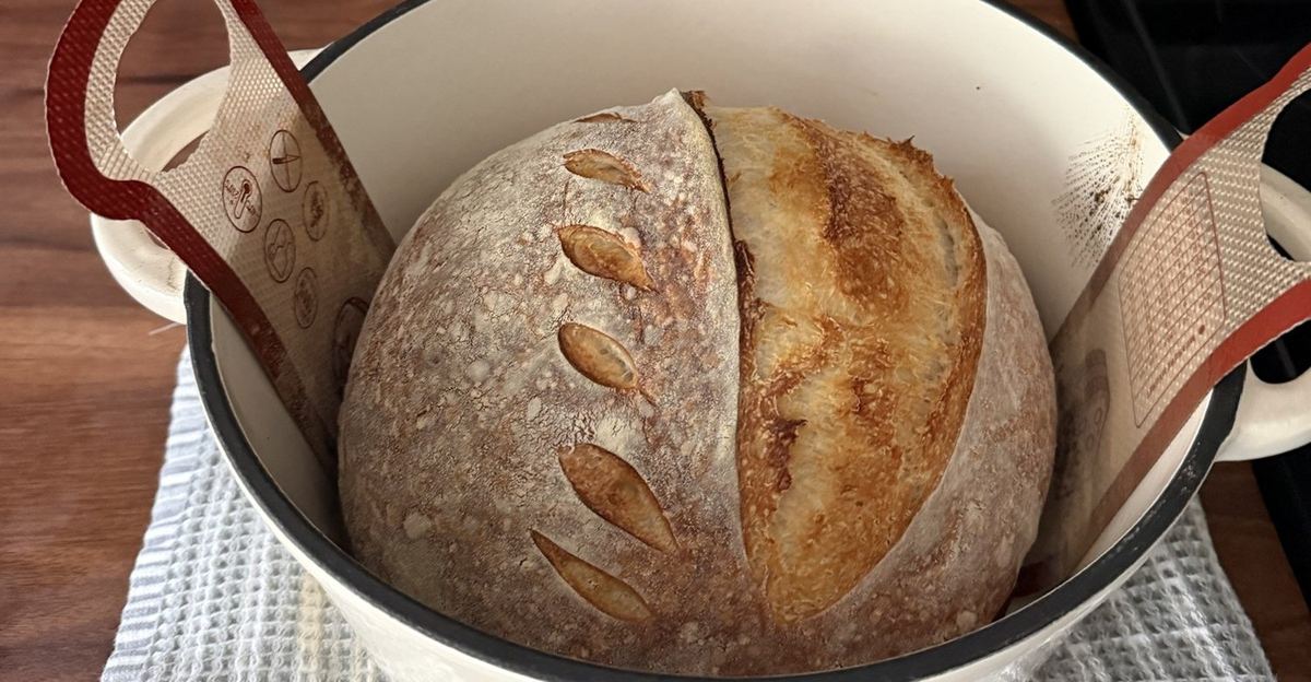 Sourdough loaf baking inside an enameled cast-iron Dutch oven, lifted by a silicone sling resting on a kitchen towel